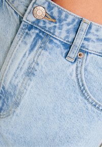 Ljusblå denimjeans med en urtvättad textur. Funktioner inkluderar en metallknapp, sydda accenter och en framficka. Slitna detaljer är närvarande.