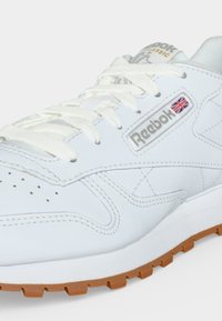 Biele kožené tenisky Reebok Classic s bielymi šnúrkami, logom Union Jack na boku a hnedou gumovou podrážkou viditeľnou na svetlom pozadí.