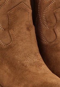Botas de cowboy de ante en color marrón claro con costuras decorativas en un patrón floral, puntera afilada y un acabado ligeramente texturizado.