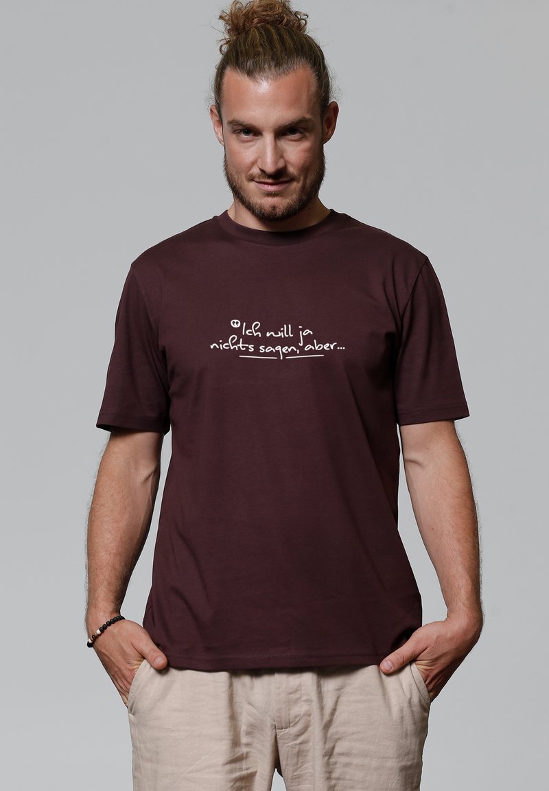 Man met lichtbruin haar in een dot draagt een donkerburgundy T-shirt met de Duitse tekst "Ich will ja nichts sagen, aber..." en beige broek met zijn handen in zijn zakken.