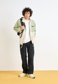 Giacca in pile verde e beige con zip, pantaloni cargo neri e sneakers chiare con dettagli. Il modello tiene uno zaino nero contro uno sfondo semplice.