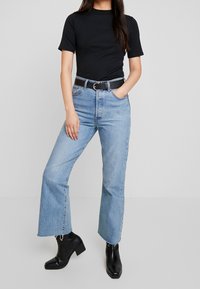 Top corto nero a maniche corte, jeans azzurri a vita alta con orlo sfilacciato, cintura nera con dettagli in argento e stivaletti neri con tacco.