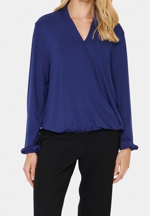 Mujer con una blusa cruzada de manga larga azul marino con cintura elástica y pantalones negros, de pie contra un fondo claro y sencillo.