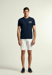 Camiseta de algodón azul marino con un estampado gráfico en el pecho, combinada con pantalones cortos blancos. El modelo está de pie sobre un fondo neutro.