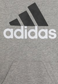 Felpa grigia con un tessuto testurizzato. Presenta un grande logo Adidas nero e una tasca frontale. Design semplice senza motivi o accenti.