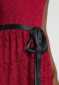 Robe en dentelle rouge ornée d'un motif floral, avec un ruban en satin noir noué à la taille, ajoutant du contraste et des détails au design.