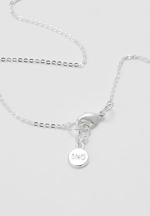 Collana a catena in argento con chiusura a moschettone e una piccola targhetta rotonda incisa con la parola "SNÖ" su uno sfondo chiaro.