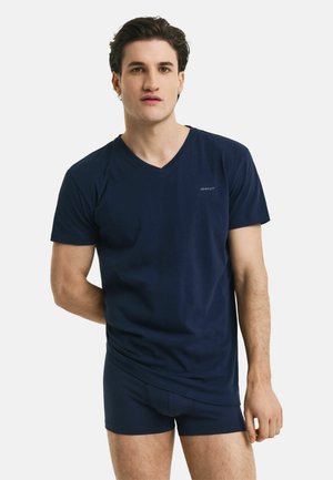 Joven con camiseta azul oscuro de cuello en V y calzoncillos bóxer a juego, de pie frente a un fondo blanco liso.