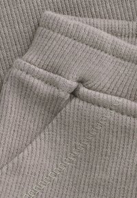 Close-up van ribbel gebreide stof in lichtgrijs met zichtbare naden en gevouwen randen, waarop de textuur en stikseldetails te zien zijn.