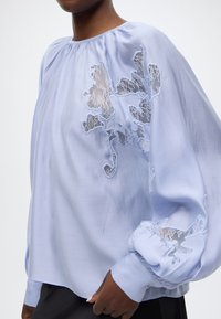 Ανοιχτό μπλε μπλούζα με στρογγυλή λαιμόκοψη, μακριά φουσκωτά μανίκια και περίτεχνη δανδέλα κεντήματα στο μπροστινό μέρος και τις μανσέτες. Λείο υφασμάτινο υφή.
