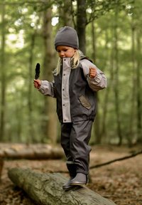 Veste imperméable grise avec accents gris clair, pantalon assorti et bottes grises, portée par un enfant tenant une plume debout sur une bûche.