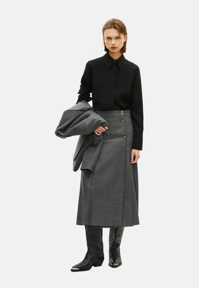 Chemise noire à col accompagnée d'une longue jupe grise à carreaux avec des boutons dorés. Un blazer gris est drapé sur le bras, complété par des bottes noires.