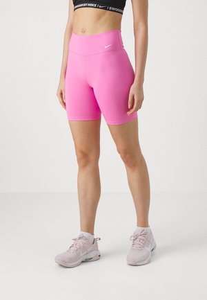 Femme portant un short de vélo Nike taille haute rose vif, un soutien-gorge de sport noir Nike et des baskets Nike rose clair, debout devant un fond uni.