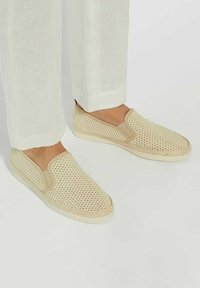 Beige slip-on shoes with a woven mesh design, contrasting tan accents, and a thick white outsole, paired with light linen pants.