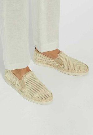 Beige slip-on shoes with a woven mesh design, contrasting tan accents, and a thick white outsole, paired with light linen pants.