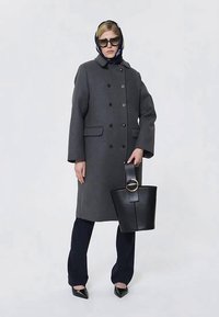 Kvinna klädd i en mörkgrå dubbelknäppt kappa, svarta byxor, scarf, solglasögon och håller en svart läderhandväska med stort runt spänne.