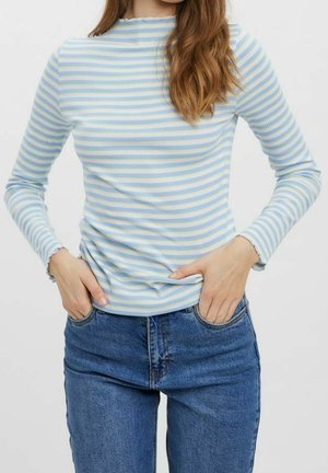 Vrouw met een lichtblauw-wit gestreept shirt met lange mouwen en kroesrandjes, ingestopt in een hooggetailleerde blauwe spijkerbroek, handen in de voorzakken.