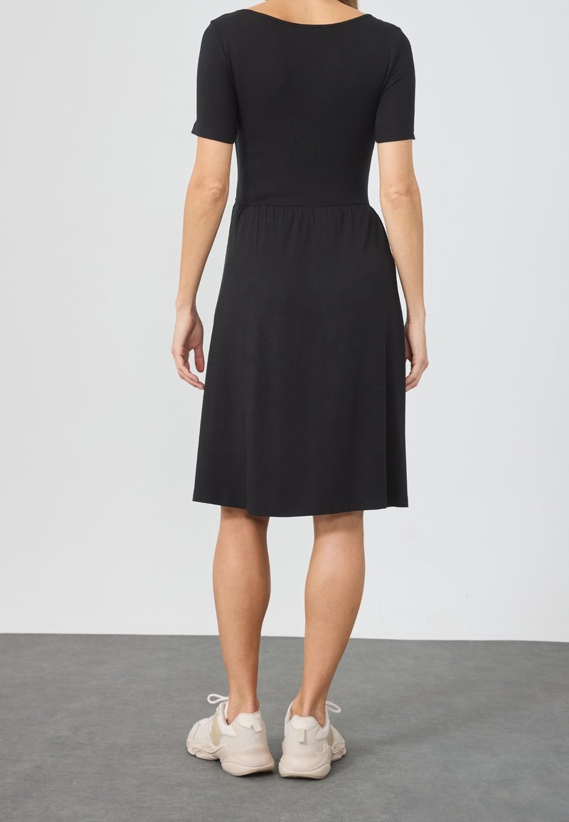 Černé krátké šaty s přiléhavým živůtkem a rozšířenou sukní. Hladká látka, délka po kolena, nošeny s světle zbarvenými teniskami.