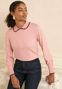 Hellrosa Pullover mit rundem Kragen, verziert mit einem rand aus dunklen Pompons. Lange Ärmel, figurbetonte Passform, kombiniert mit hochtaillierten Jeans aus dunklem Denim.