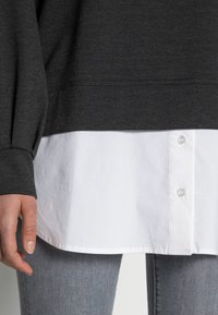 Felpa grigia scura con tessuto testurizzato sovrapposta a una maglietta bianca, con orlo dritto e bottoni visibili. Sono indossati jeans grigi.