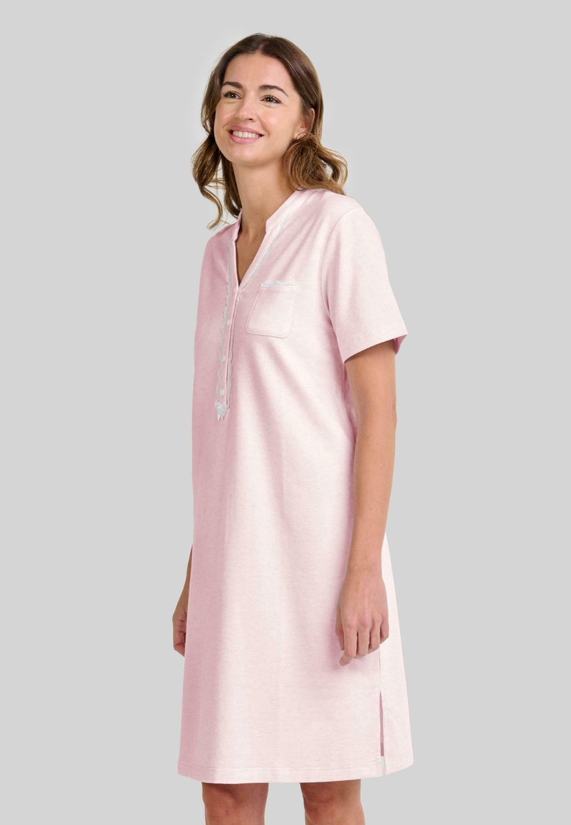 Mujer sonriente vestida con un vestido de manga corta, de longitud a la rodilla y color rosa claro, con botones en la parte frontal y un pequeño bolsillo en el pecho, de pie frente a un fondo gris.