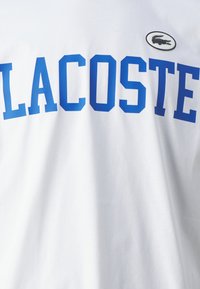 Balta medvilninė marškinėliai su ryškiu mėlynu "LACOSTE" užrašu ir mažu juodu krokodilu ant balto fono. Trumpomis rankovėmis.