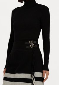 Svart ribbad polotröja som kombineras med en svart kjol med breda grå ränder, två svarta remmar med guldfärgad spänne och fransdetaljer.