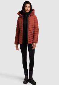 Paksustatud, roostekarva jope kõrge kaelusega ja reguleeritava kapuutsiga. Omadusteks vertikaalsed õmblused, tõmblukuga taskud ja mustad detailid.