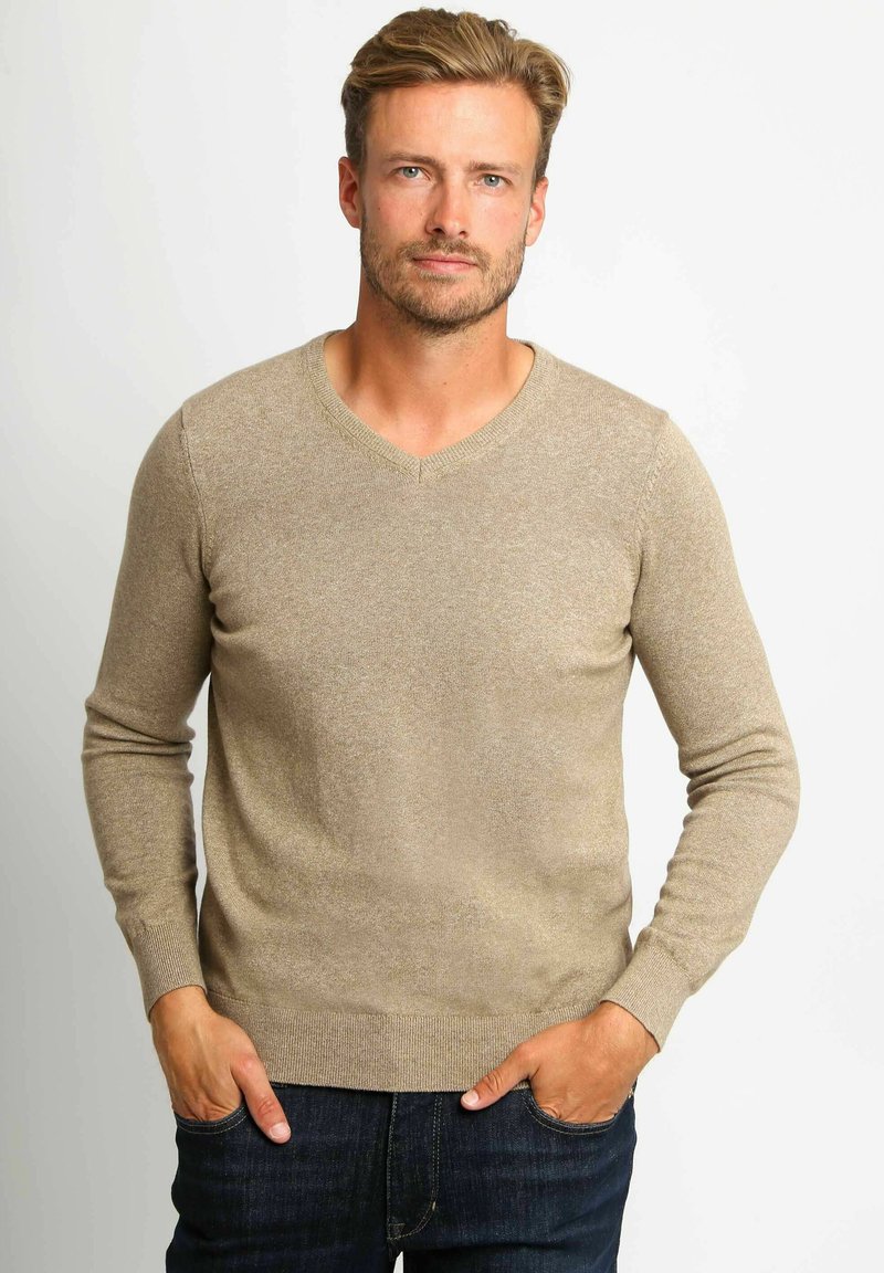 Beige gebreide trui met een V-hals, lange mouwen en geribbelde manchetten. Draagt over donkerblauwe jeans, de stof heeft een subtiele textuur.