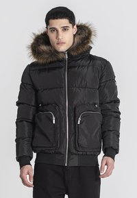 Giubbotto imbottito nero con cappuccio foderato in pelliccia sintetica, due tasche frontali con zip e polsini a costine. Materiale liscio e lucido, design trapuntato.