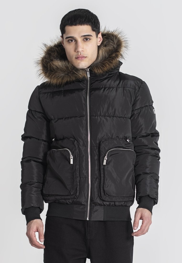 Giubbotto imbottito nero con cappuccio foderato in pelliccia sintetica, due tasche frontali con zip e polsini a costine. Materiale liscio e lucido, design trapuntato.