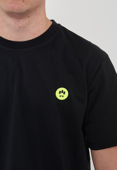 T-shirt en coton noir avec un petit graphique rond de visage néon jaune avec des crocs sur la partie gauche de la poitrine. Manches courtes et col rond.