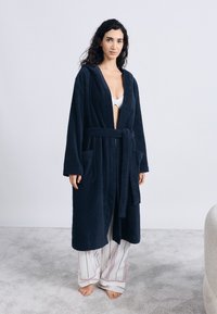 Μπλε ναυτικό μπουρνούζι από περκάλι με κουκούλα, ζώνη δέσιμο και δύο μπροστινές τσέπες. Φορεμένο πάνω από άσπρο μπουστάκι και ριγέ πιτζάμες.