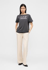Szara koszulka z krótkim rękawem z logo "EA7 EMPORIO ARMANI", zestawiona z luźnymi kremowymi spodniami i czarnymi butami. Prosty, czysty design.