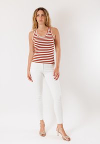 Top con righe rosse e bianche, design aderente, abbinato a jeans skinny bianchi e sandali dorati con cinturini, evidenziando un look casual.
