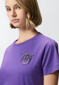 Ljubičasta majica kratkih rukava s dekorativnim perlicama u nijansama crne i ljubičaste boje na prsima, izrađena od mekanog pamučnog materijala.