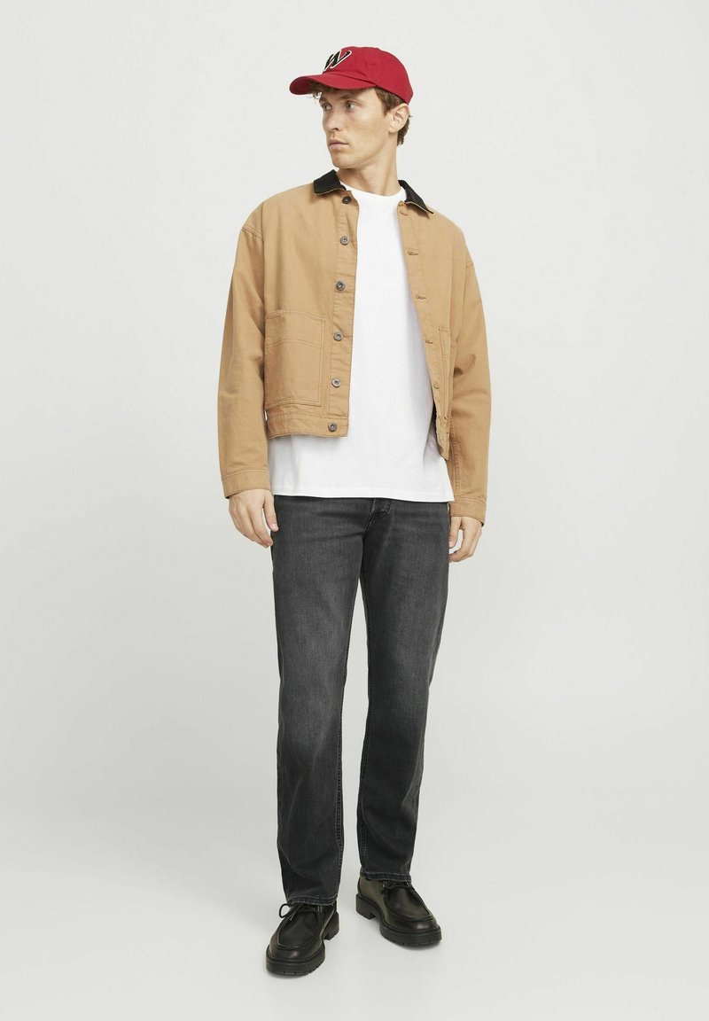 Braune gekürzte Jacke mit schwarzem Kragen, weißem T-Shirt, dunkelgrauen Jeans und rotem Baseballcap. Schwarze Schuhe runden das Outfit ab.