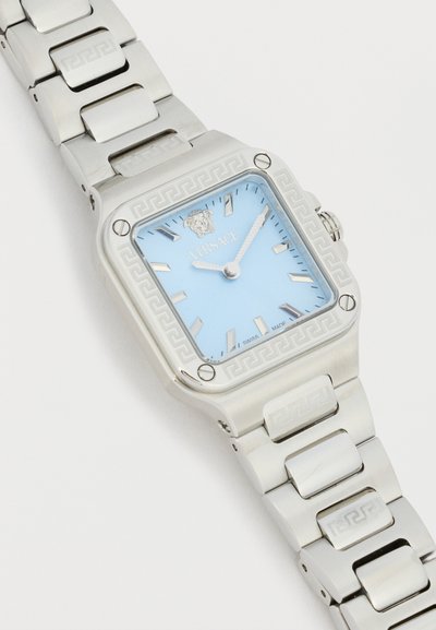 Ceas de mână pătrat argintiu cu cadran albastru deschis, marcatoare argintii, logo Medusa la ora 12 și brățară metalică cu modele gravate.