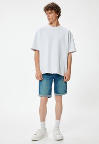 Camisa blanca de gran tamaño con dobladillo sin acabar, combinada con pantalones cortos de denim y zapatillas blancas. Tejido suave, ajuste casual. Puños enrollados en los pantalones cortos.
