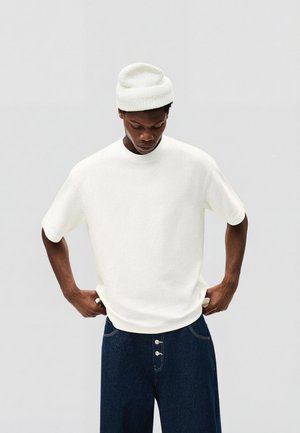 Man met witte gebreide muts en wit shirt met korte mouwen, die een donderblauwe spijkerbroek met knoopsluiting aanpast, tegen een effen lichte achtergrond.