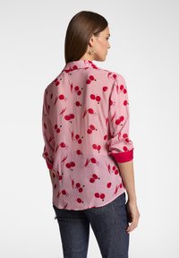 Blusa rosa chiaro con motivo floreale rosso, maniche arrotolate e polsini rossi solidi. Tessuto morbido, vestibilità rilassata e orlo curvo.