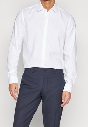 Homme portant une chemise blanche à manches longues boutonnée, rentrée dans un pantalon de costume bleu marine foncé, sur un fond uni.