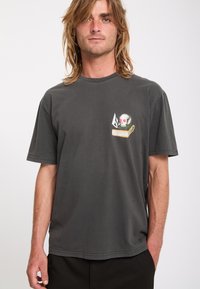 Jeune homme aux cheveux blonds mi-longs portant un t-shirt gris foncé avec un motif de crâne et de livre sur la poitrine.