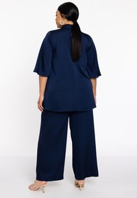 Marineblauwe, losse top met een hoge kraag en klokmouwen, gecombineerd met broek met wijde pernas. Gladde textuur, minimalistisch ontwerp en subtiele flow.
