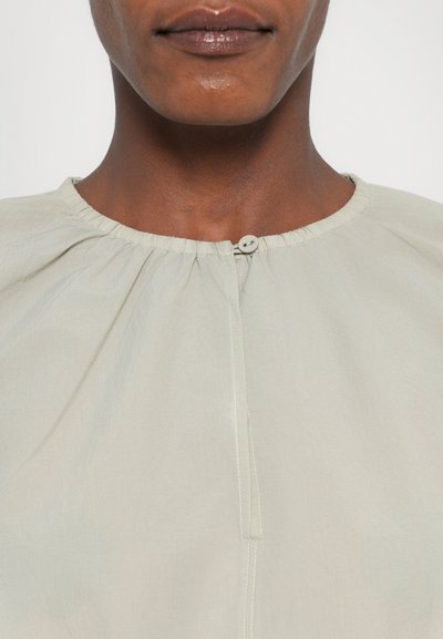 Blusa verde chiaro con scollo arricciato, chiusura frontale con bottoni e dettagli di cucitura sottili. Tessuto liscio con una vestibilità rilassata.