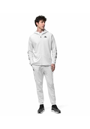 Jeune homme debout portant un sweat à capuche blanc zippé et un pantalon de jogging assorti avec un logo noir, associé à des baskets noires et blanches.
