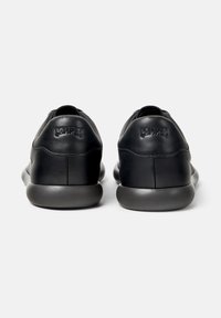 Pair of black leather sneakers with rounded dark gray soles viewed from the back, featuring embossed "Camper" branding on the heels.