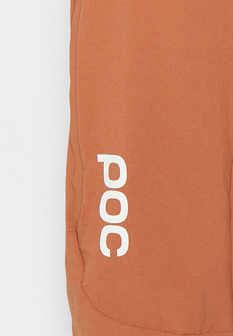 Jogger akin na kolorze rdzy o gładkiej teksturze, z białym logo "POC" po boku oraz detalem bocznej kieszeni.