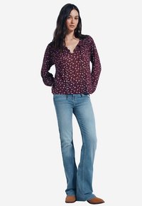 Weinrote Bluse mit Blumenmuster, V-Ausschnitt und Kordelzug, kombiniert mit hellblauen Schlagjeans. Weicher Stoff mit legerer Passform.