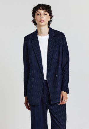 Blazer a righe navy con due bottoni, rever a punta e una tasca sul petto. Tessuto liscio, abbinato a una maglietta bianca.